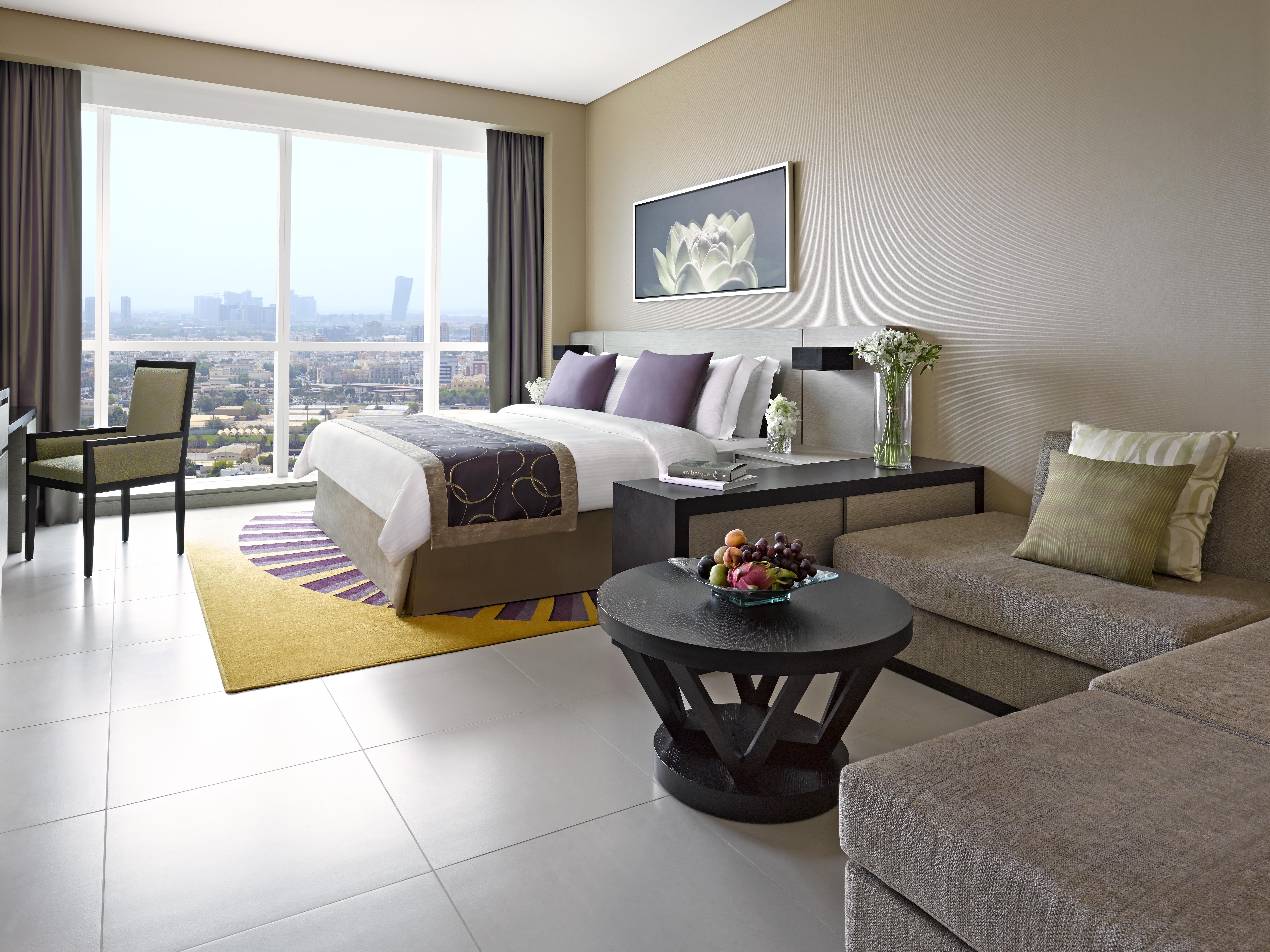 Dusit Thani Абу-Даби Экстерьер фото The photo shows a modern hotel room featuring a combination of a bedroom and a living area. There is a large bed with white linens and decorative purple pillows, positioned against a wall. Next to the bed, there is a bedside table and a small vase wi
