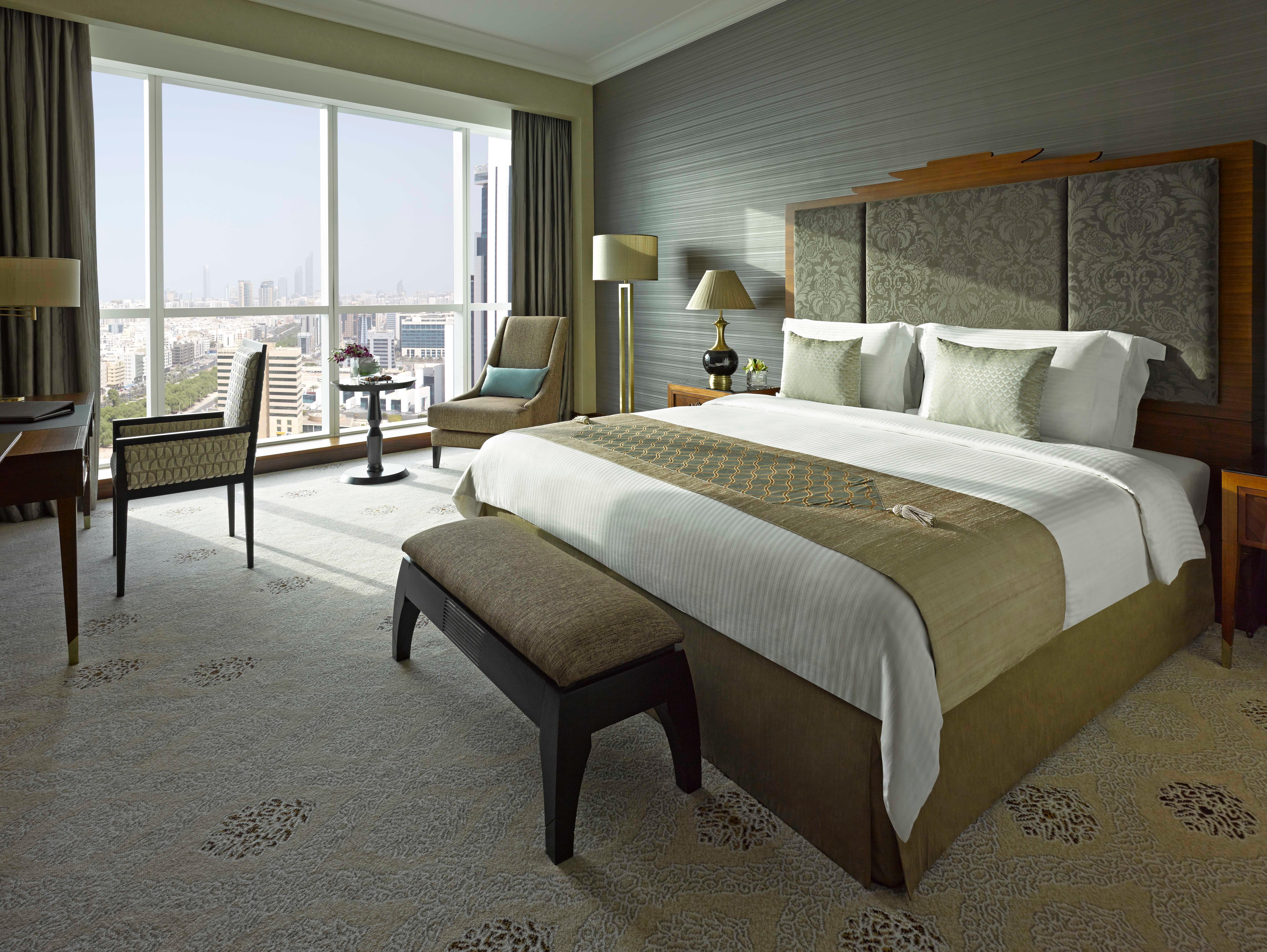 Dusit Thani Абу-Даби Экстерьер фото The photo shows an elegantly designed hotel room. It features a large king-sized bed with white linens and a beige decorative cover. Flanking the bed are bedside tables with lamps. 

In the background, there's a large window that offers a view of the