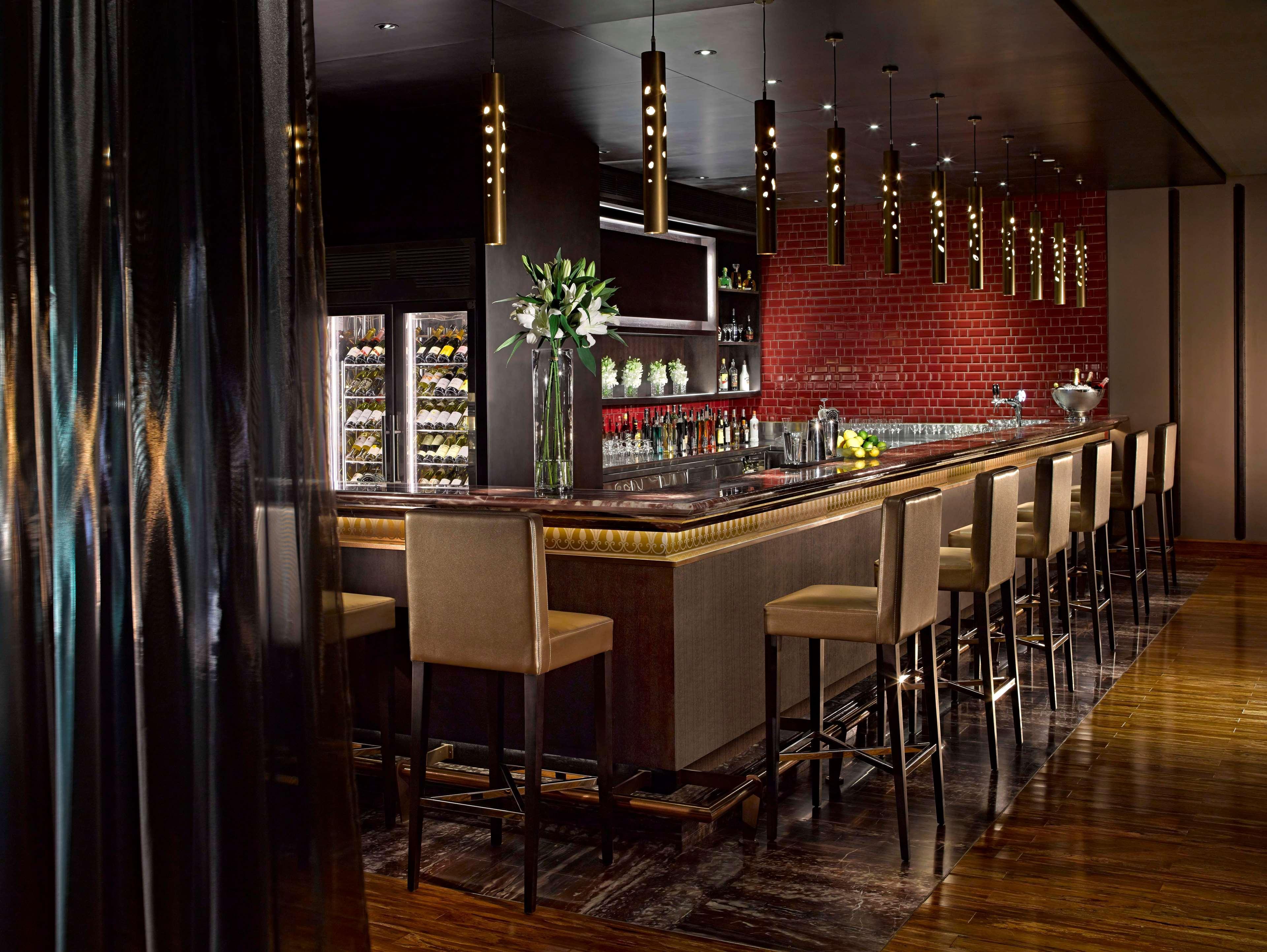 Dusit Thani Абу-Даби Экстерьер фото The photo depicts a stylish and modern bar area. There is a long bar countertop with a sleek design, featuring elegant lighting fixtures hanging above it. The bar is lined with high stools, each with a light-colored upholstery. Behind the bar, there 