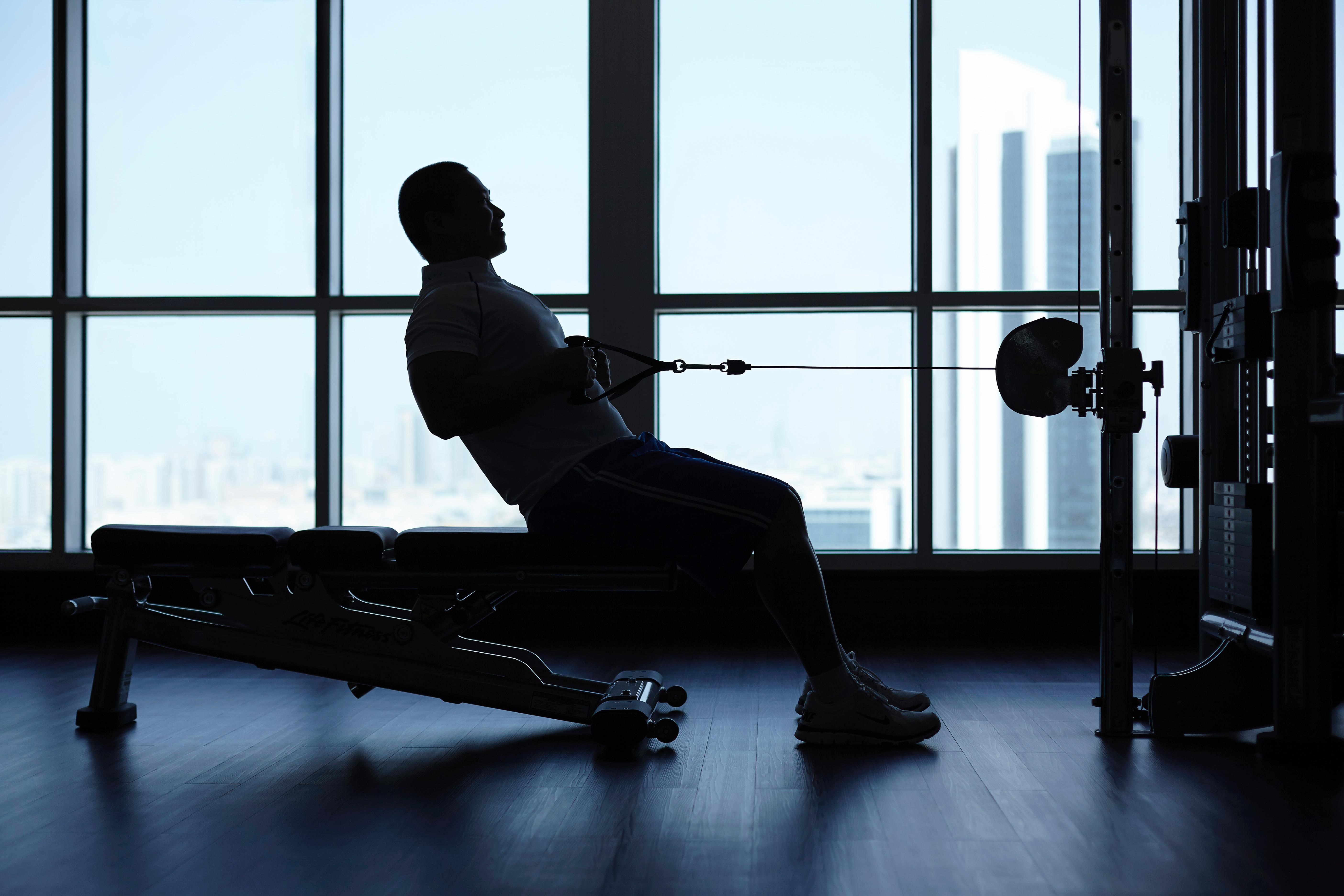 Dusit Thani Абу-Даби Экстерьер фото The photo shows a silhouette of a person exercising in a gym. The individual is seated on a bench, using a cable machine, and appears to be performing a pulling exercise. The backdrop features large windows that provide a view of a cityscape, with ta