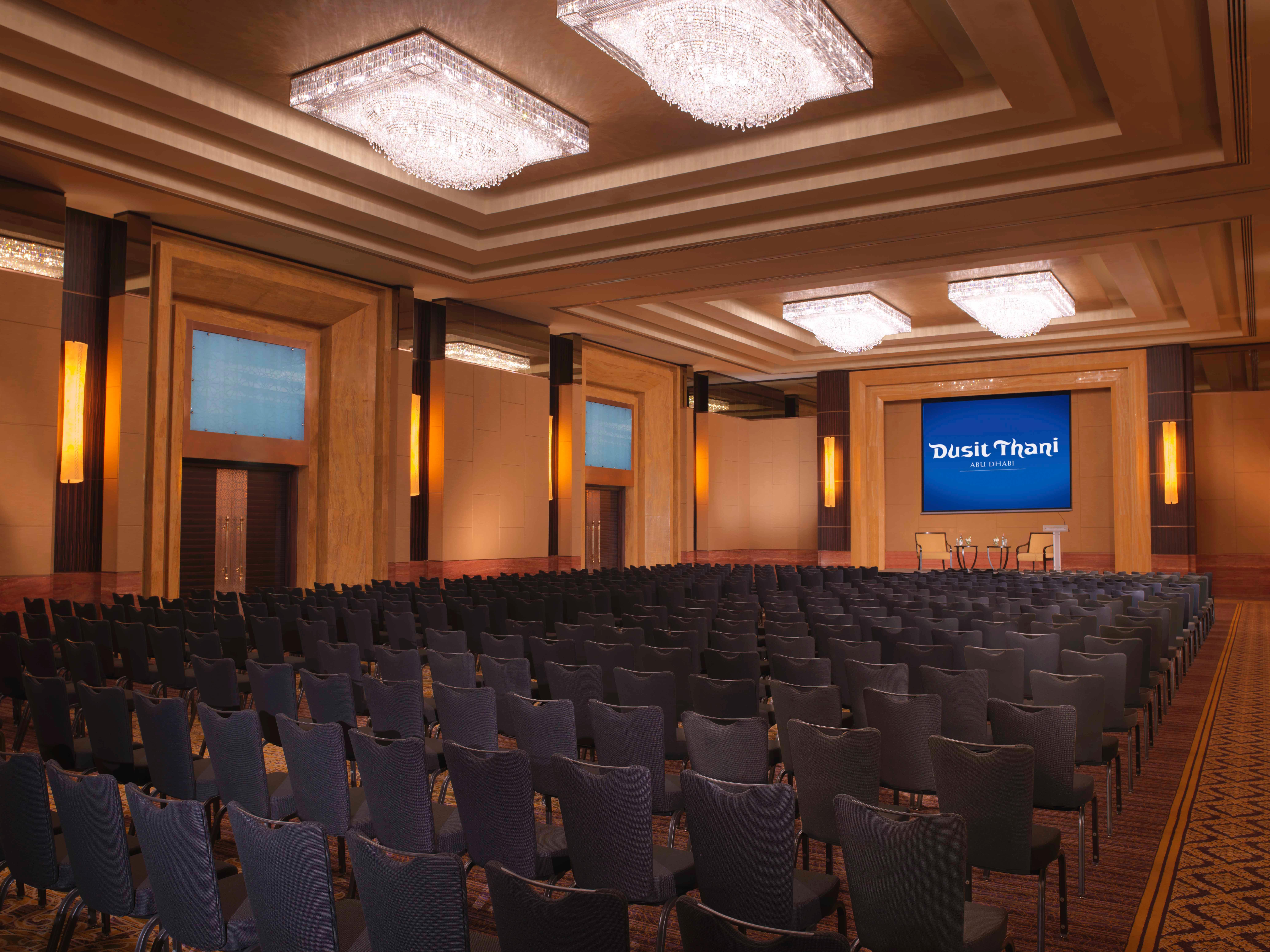 Dusit Thani Абу-Даби Экстерьер фото The photo shows a well-lit auditorium or conference room set up for an event. The space features a large number of dark chairs arranged in rows facing a stage or presentation area. Above the stage, there is a screen displaying the text "Thank You!" i