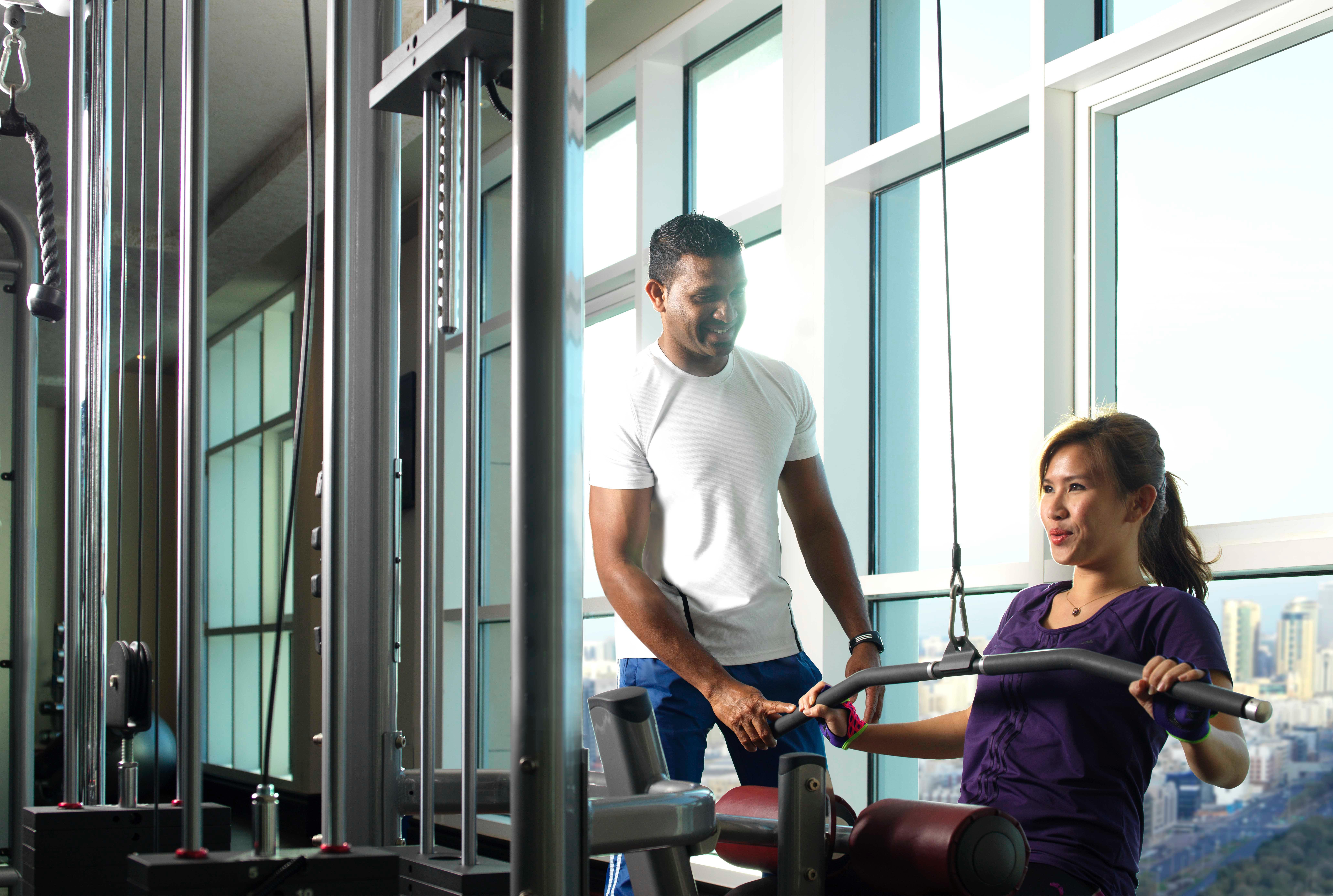 Dusit Thani Абу-Даби Экстерьер фото The photo shows two individuals in a gym setting. One person, likely a trainer, is assisting a woman who appears to be exercising on a machine. The gym has large windows that let in natural light, creating a bright atmosphere. The trainer is focused 