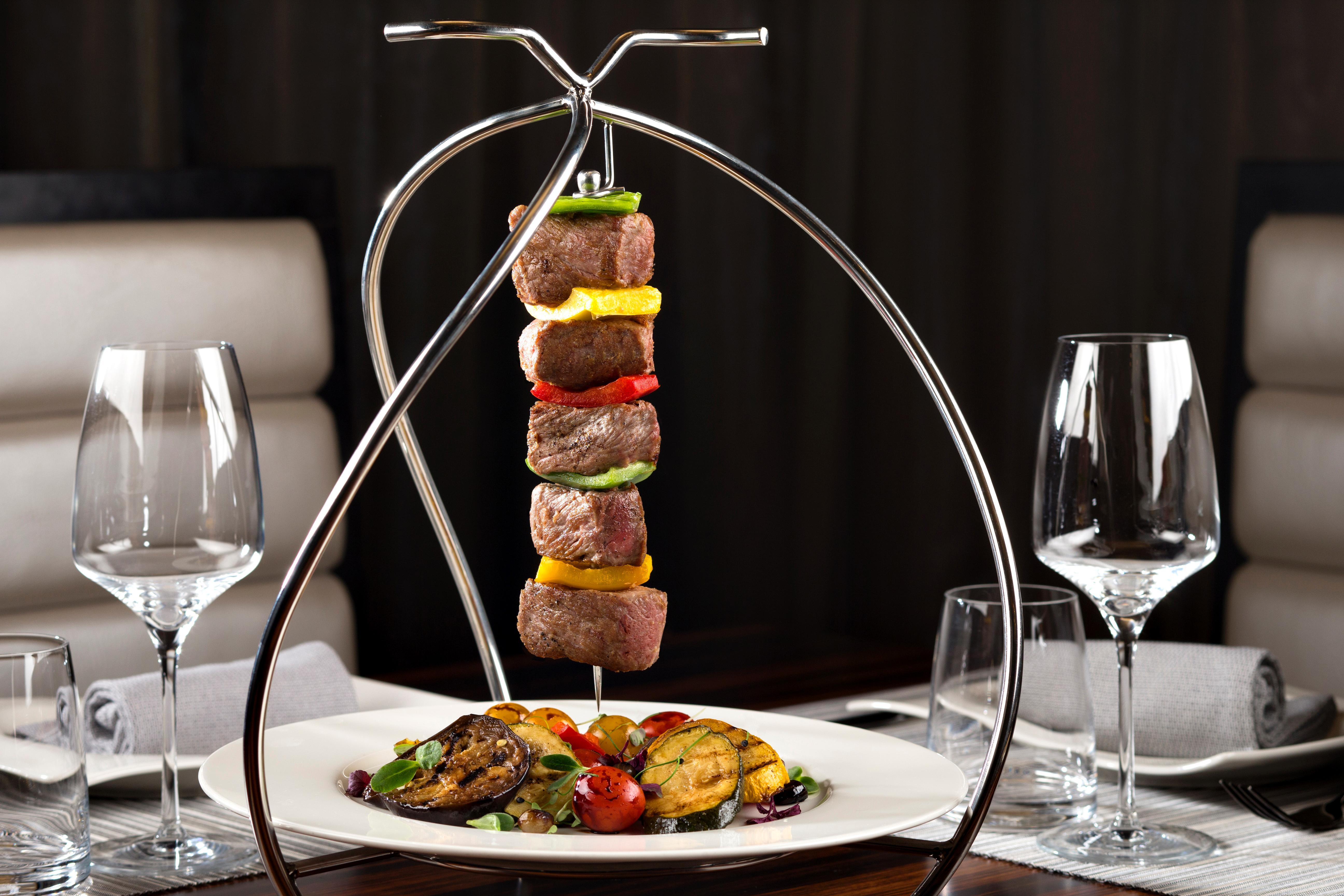 Dusit Thani Абу-Даби Экстерьер фото The photo shows a stylish presentation of skewered meat and vegetables. The skewers are hanging from a metallic stand, featuring chunks of beef interspersed with colorful bell peppers and onions. Below the skewers, there is a plate that includes gril