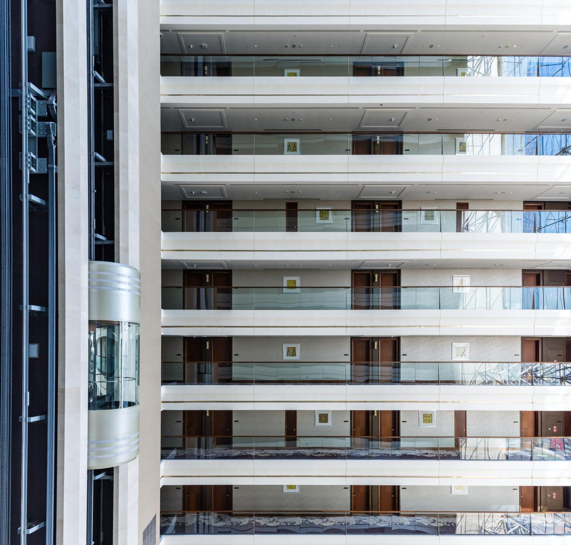 Dusit Thani Абу-Даби Экстерьер фото The image shows the interior of a multi-story building, likely a hotel or modern residential structure. It features several floors with balconies that have glass railings. The walls are finished in a light color, and there are doors leading to indivi